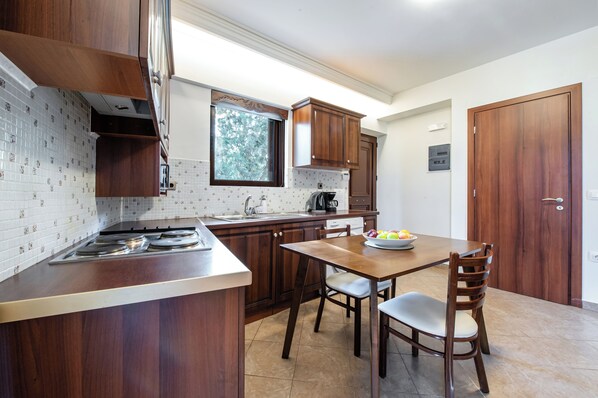A kitchen with wooden cabinets, a gas stove, a dining table with chairs, and a window. - Kahlua Bay Apartments (Hersonissos)
