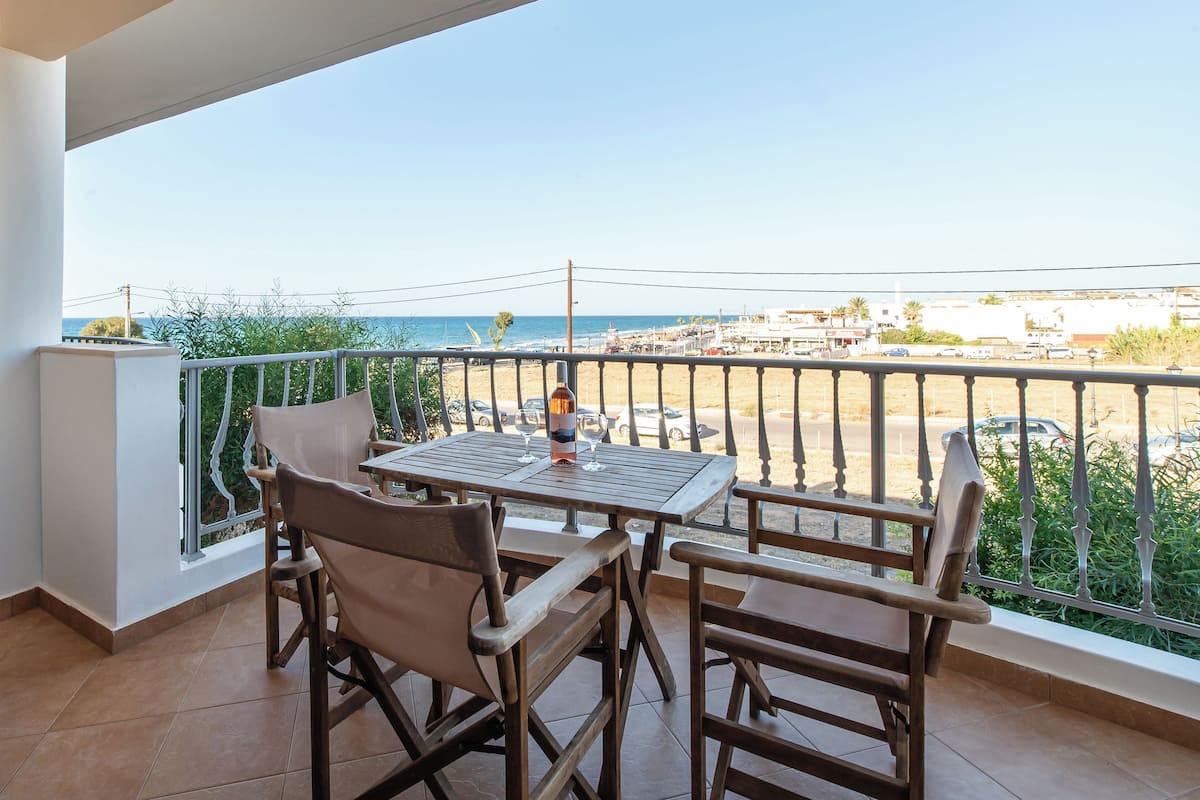 A balcony with a table and chairs, overlooking a beach and parking area.