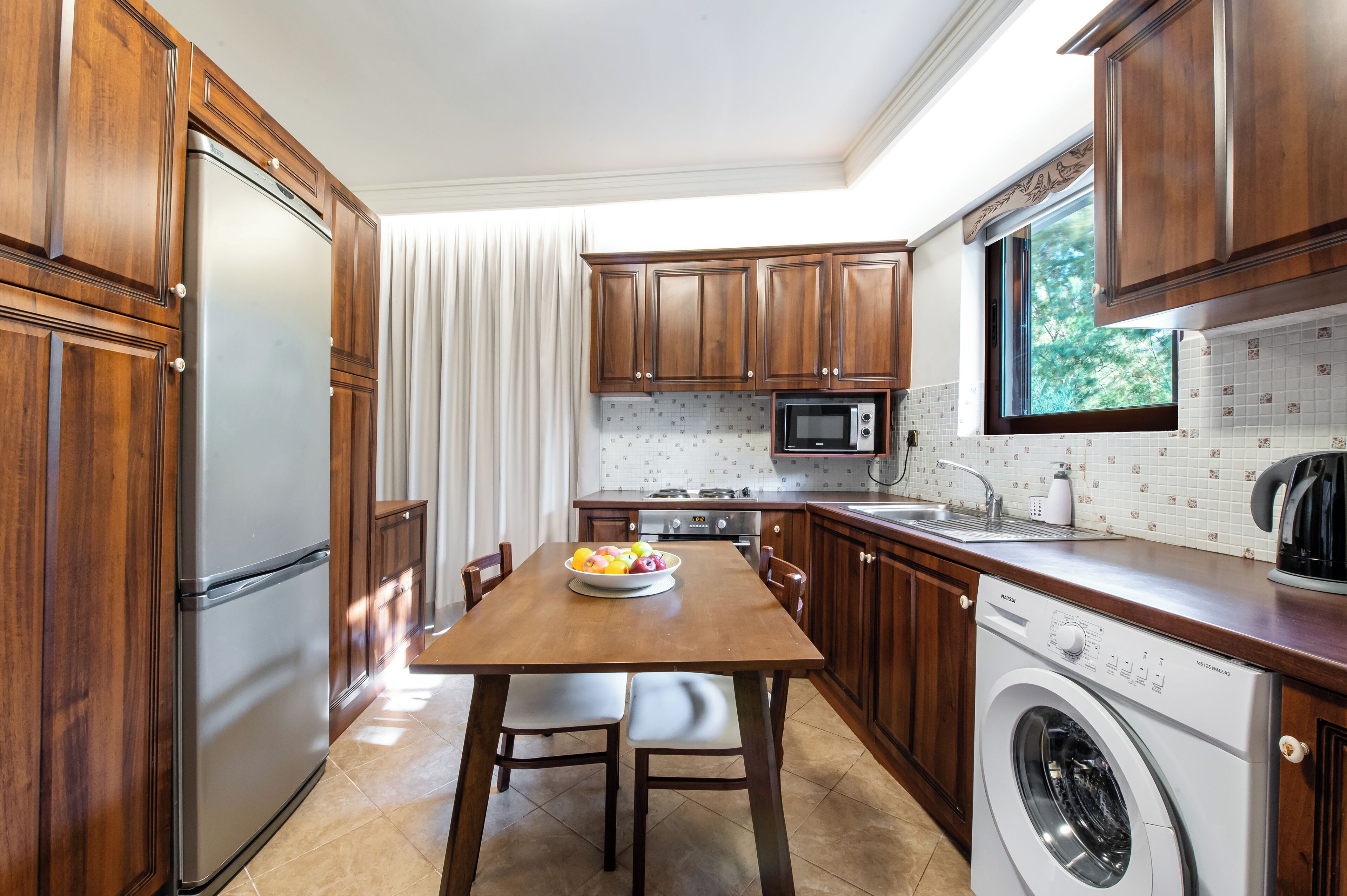 A kitchen with wooden cabinets, a stainless steel refrigerator, a microwave, a sink, and a washing machine.