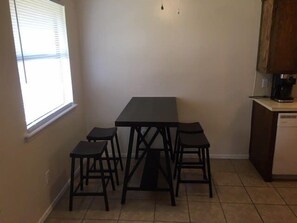 A small kitchen with a table and four stools, a window with blinds, and a coffee machine.