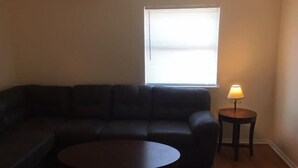 A living room with a dark-colored sofa, a round coffee table, and a lamp on a small table. - Fully furnished two bedroom apartment (Lawton)