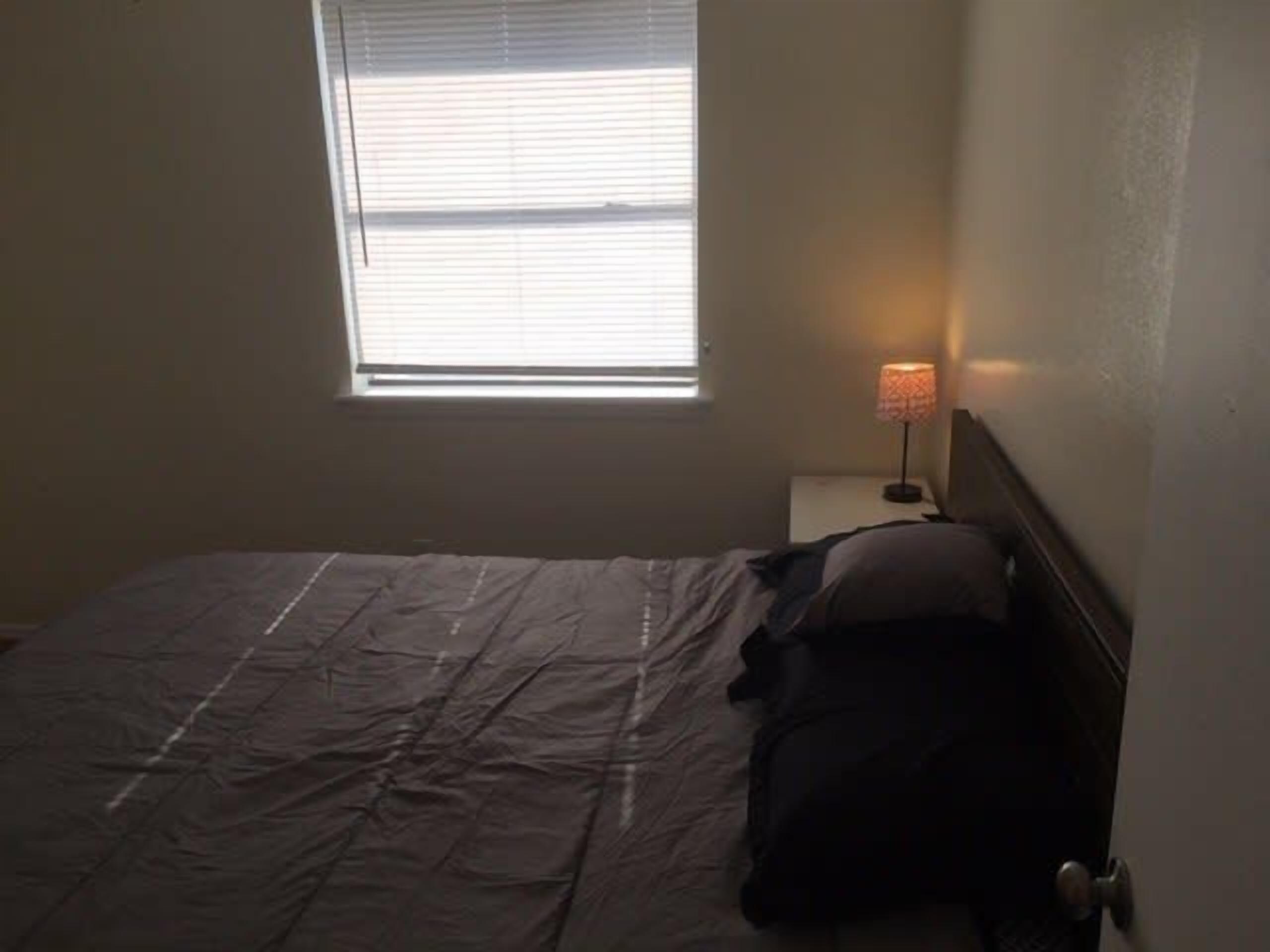 Una habitación pequeña y tenue, con una cama individual, una mesita de noche con lámpara y una ventana con persianas.