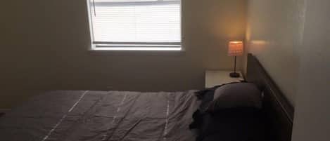 Ein kleiner, schummrig beleuchteter Raum mit einem Einzelbett, einem Nachttisch mit Lampe und einem Fenster mit Jalousien.