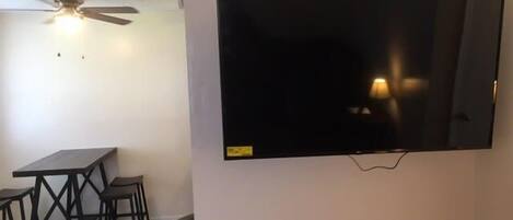 A room with a flat-screen TV mounted on the wall, a ceiling fan, a dining table with chairs, and a trash bin.