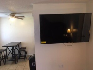 A room with a flat-screen TV mounted on the wall, a ceiling fan, a dining table with chairs, and a trash bin.