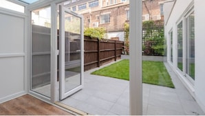 A modern room with a glass door leading to a small garden, a wooden floor, and a view of a brick building in the background. - King's Cross Garden Flat - Pass the Keys (London)