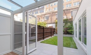 Une chambre moderne avec une porte vitrée donnant sur un petit jardin, un plancher en bois et une vue sur un bâtiment en briques en arrière-plan.