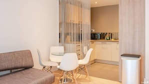 A modern living room with a sofa, a glass-top dining table, and white chairs. There is a kitchen area in the background with cabinets and appliances. - King's Cross Garden Flat - Pass the Keys (London)