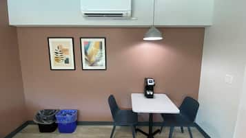 A small, modern room with a white air conditioner, two framed abstract artworks, a table with a coffee machine, two chairs, and two trash bins.