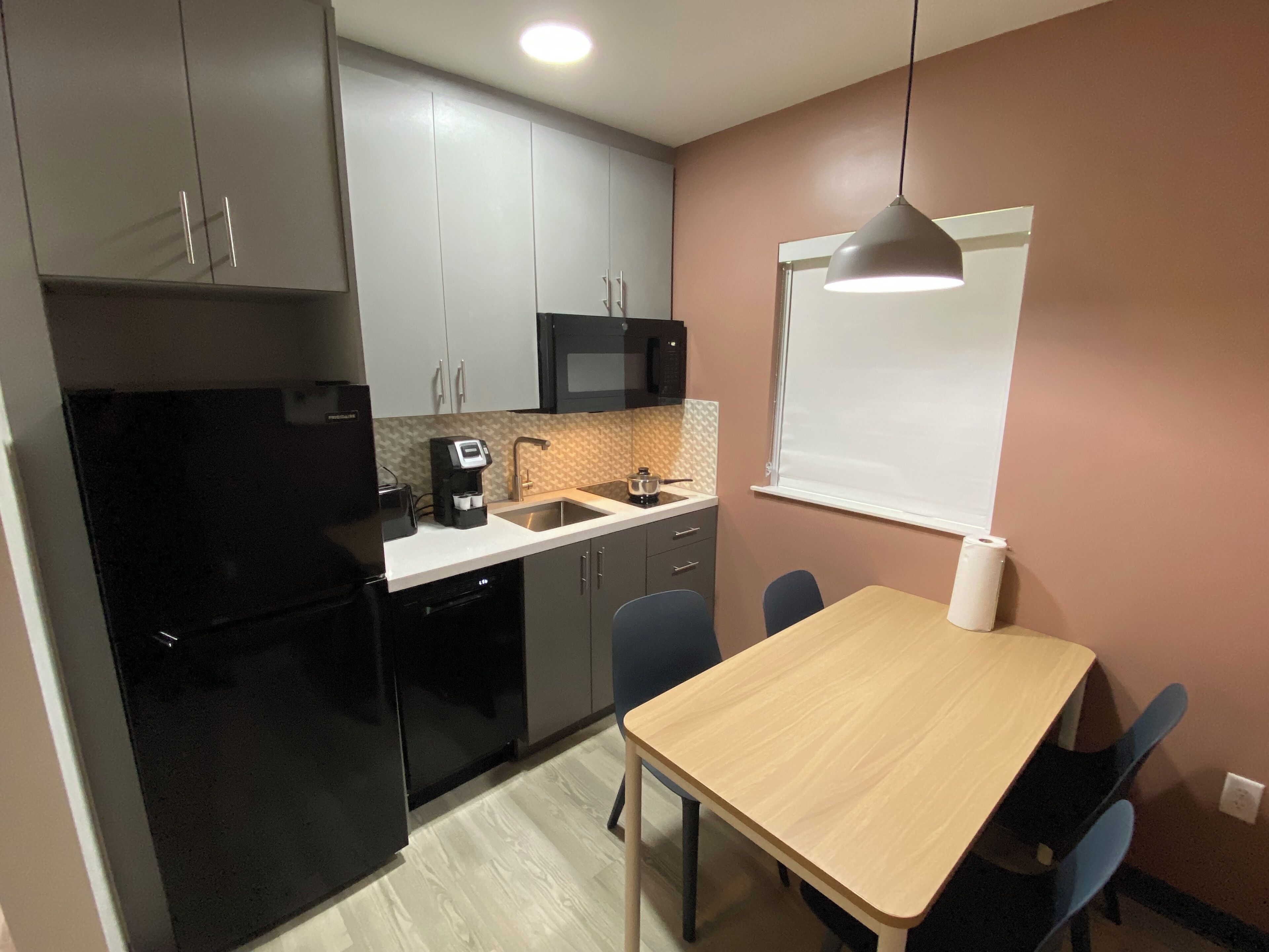 Cozinha compacta com micro-ondas, cafeteira e pia. Há uma mesa de jantar com cadeiras e um quadro branco na parede.