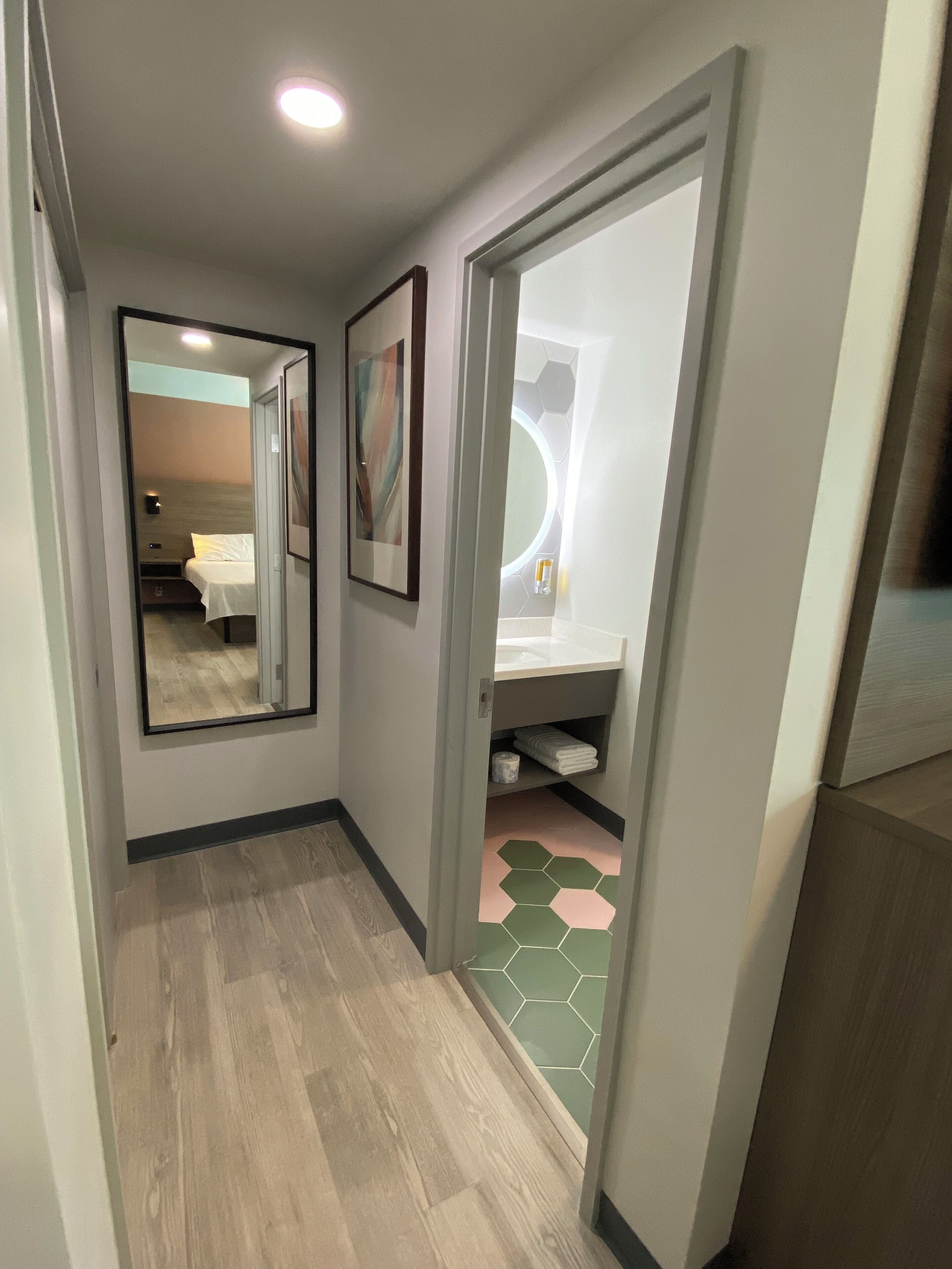 A hallway with a wooden floor, a bed with a white mattress and a headboard, a wall-mounted mirror, and a door leading to a bathroom with a green and white tiled floor.