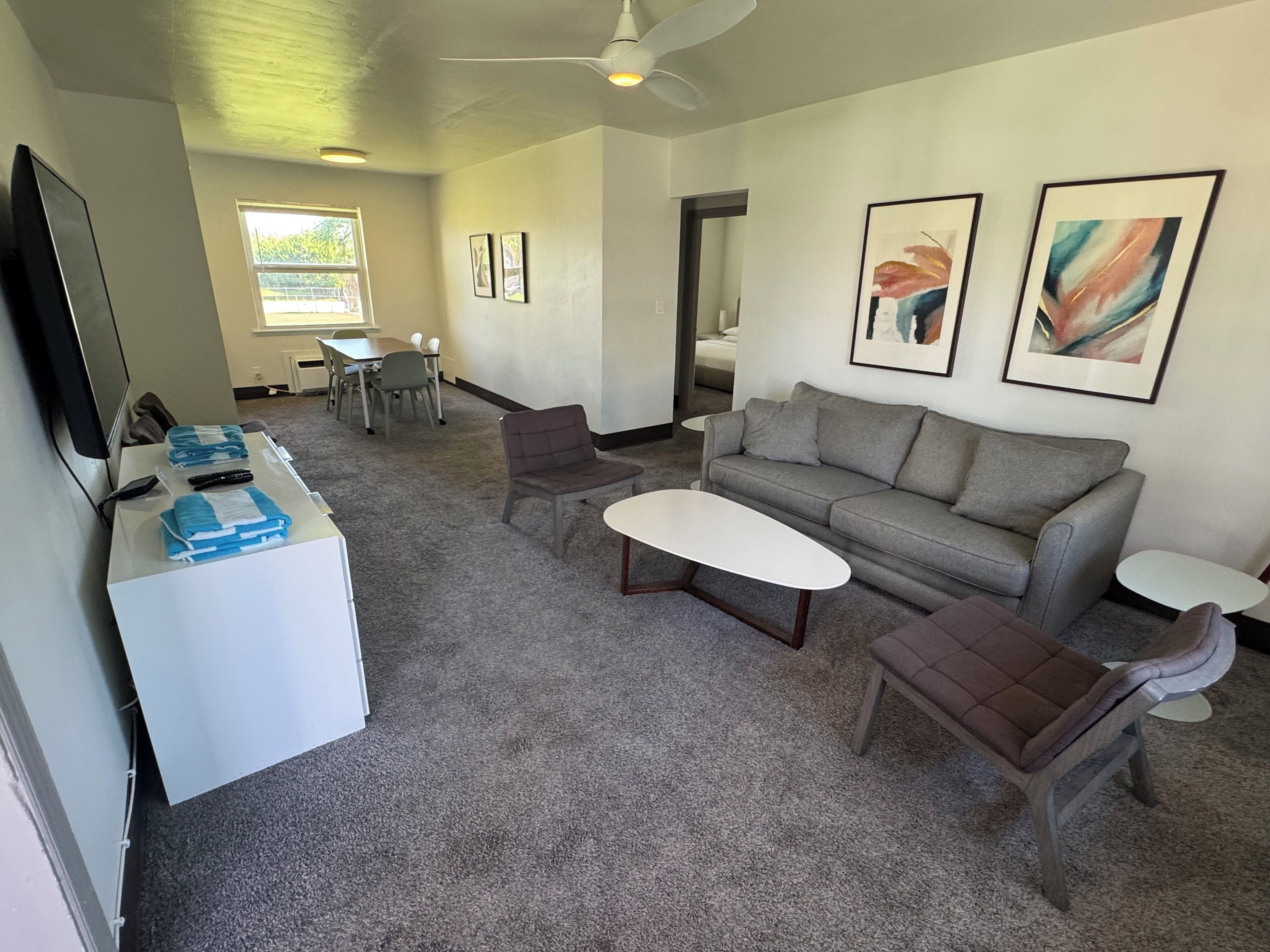 Una sala de estar moderna con un sofá gris, dos sillones, una mesa de centro y un televisor. Hay dos obras de arte enmarcadas en la pared, una mesa de comedor con sillas y una ventana con persianas.