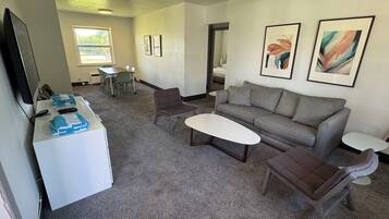 A modern living room with a grey sofa, two armchairs, a coffee table, and a TV. There are two framed artworks on the wall, a dining table with chairs, and a window with blinds.