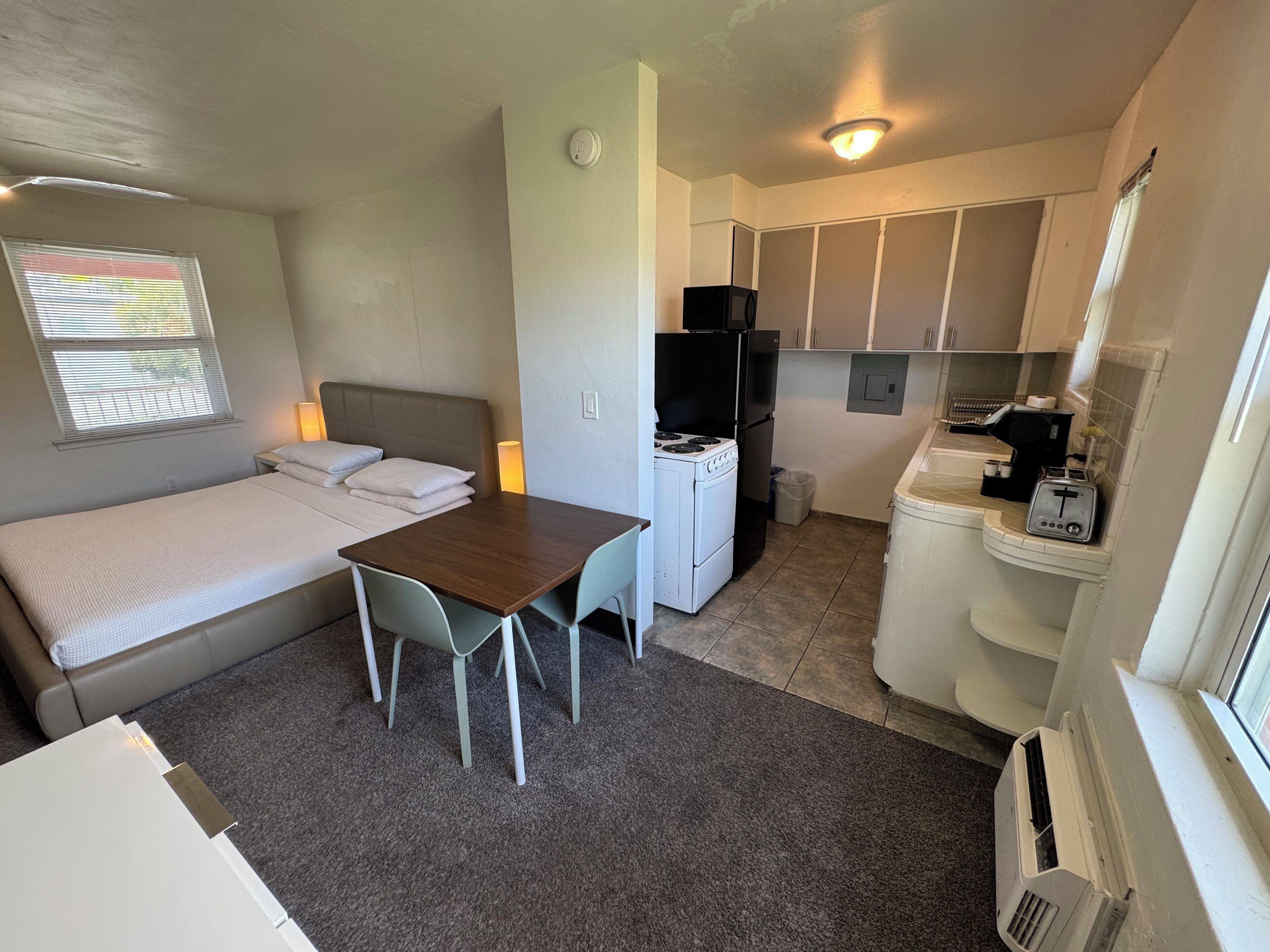 Una habitación pequeña con cama, una mesita, kitchenette y ventana con persianas.