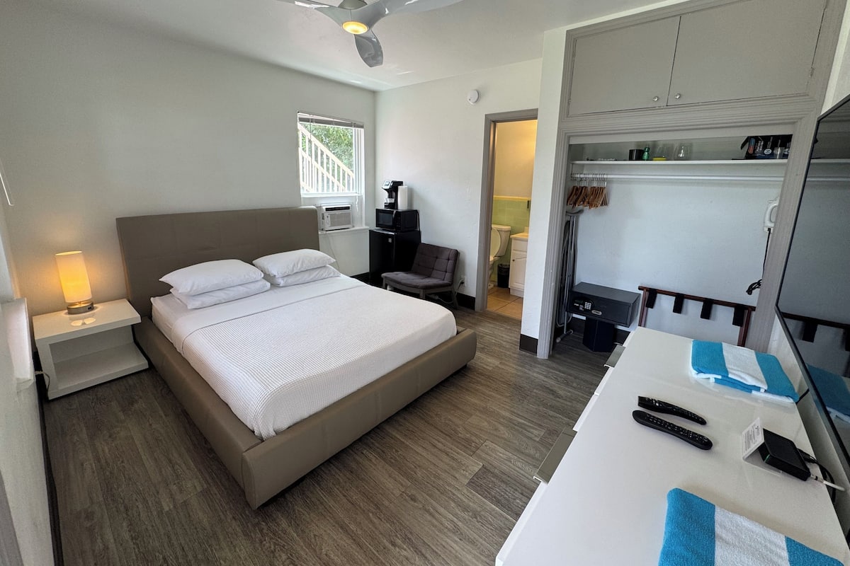 A hotel room with a bed, a desk with a computer, a chair, a ceiling fan, and a window.