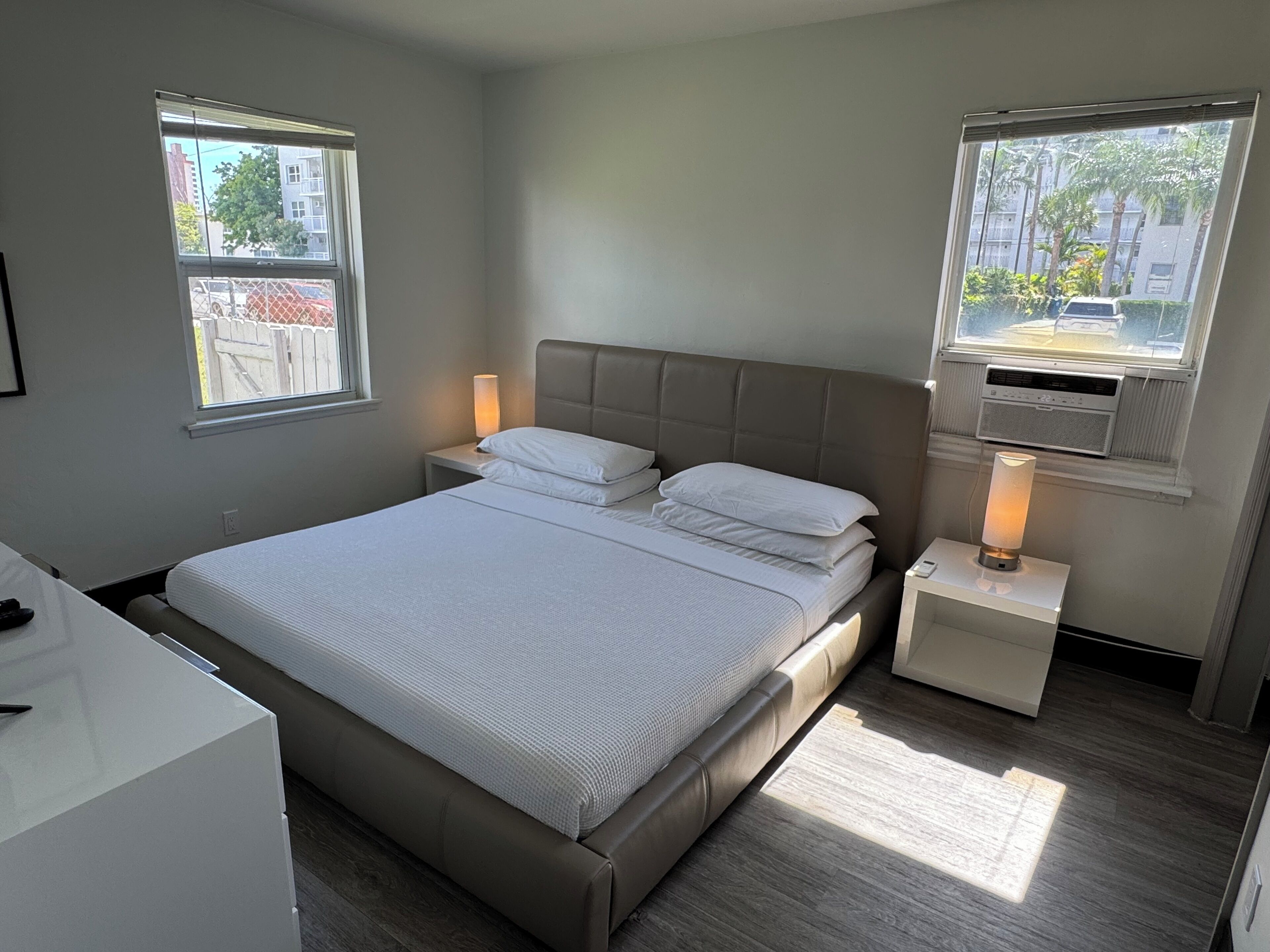 Une chambre moderne avec un grand lit, deux tables de chevet avec des lampes et des fenêtres donnant sur des immeubles et des arbres.