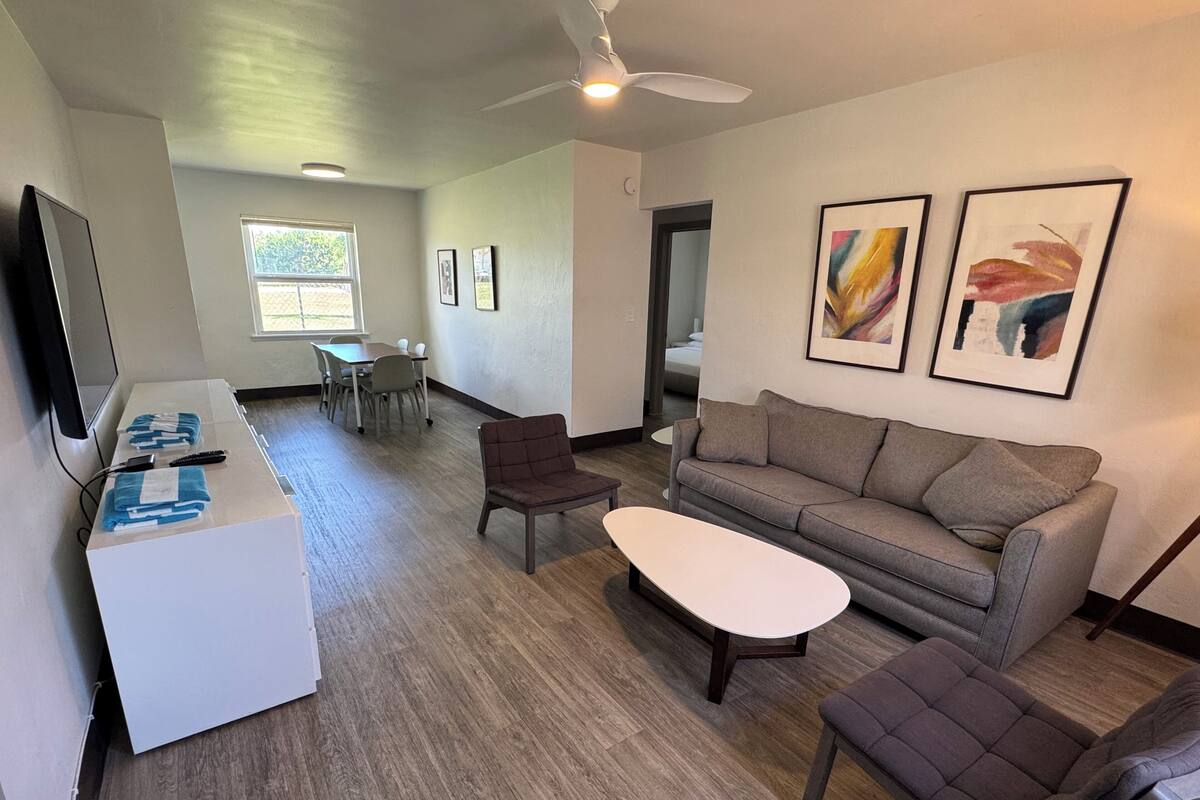 A modern living room with a grey sofa, two armchairs, a coffee table, and a TV on a stand. There is a dining area with a table and chairs, and a ceiling fan.