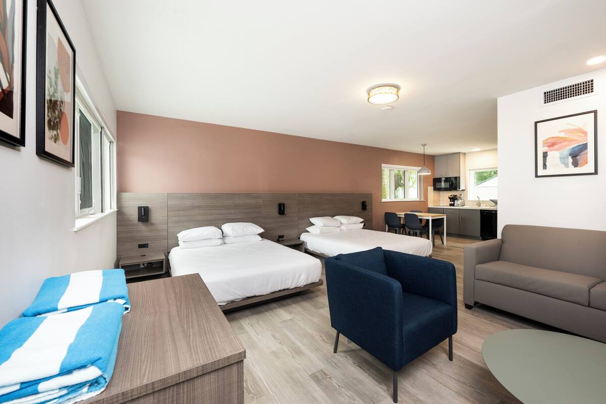 A hotel room with two beds, a sofa, a chair, a table, and a kitchenette in the background.