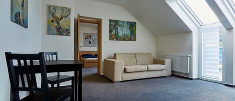 A spacious room with a high ceiling, a dining table with chairs, a sofa, and two large windows providing natural light.