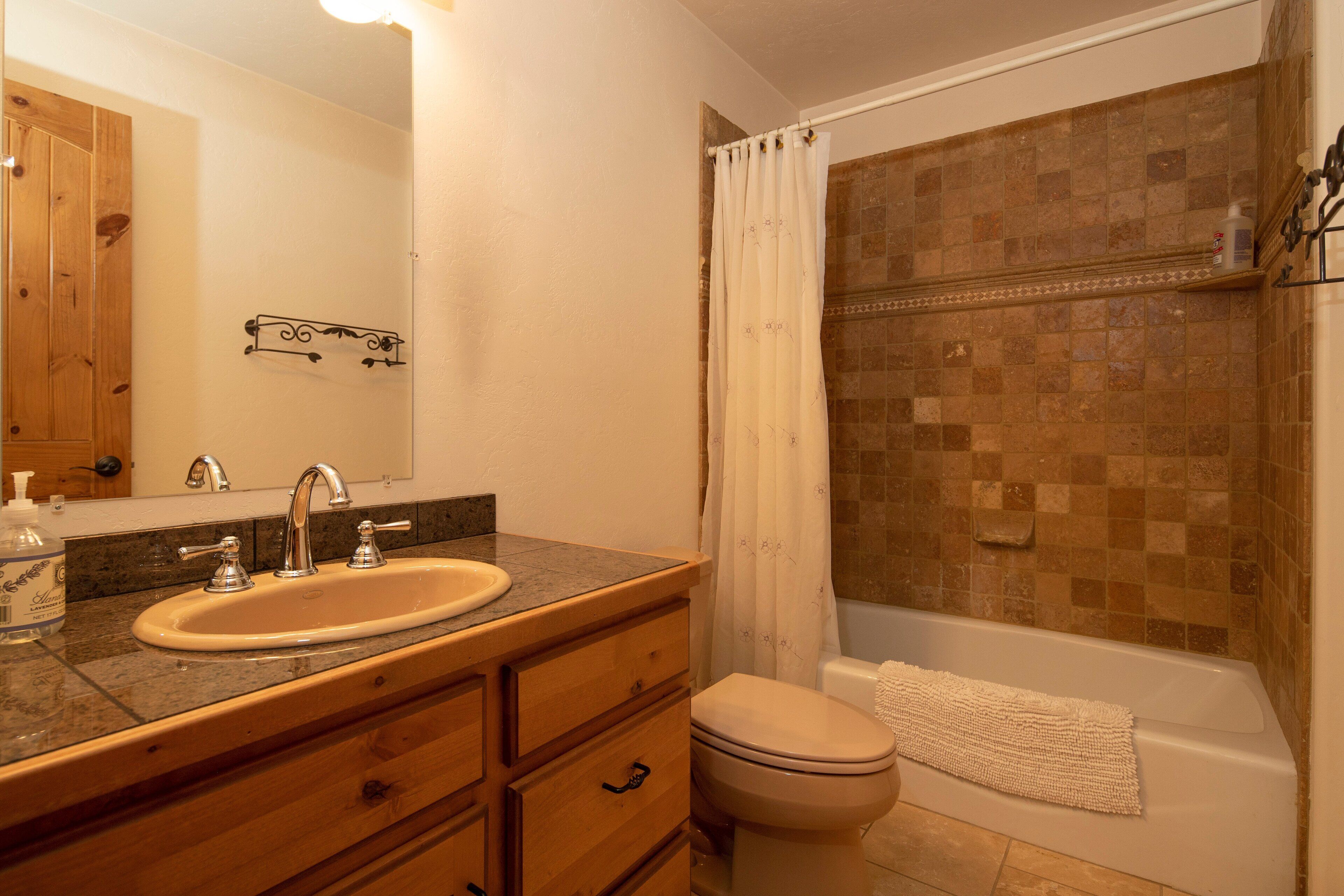 Un baño con lavabo, espejo, tocador de madera, inodoro y zona de ducha con pared de azulejos.