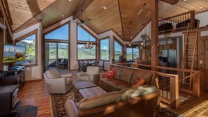 A spacious living area with a high ceiling, large windows, a cozy seating area, and a kitchen visible through an open door. - Alpine Recharge (Alpine)