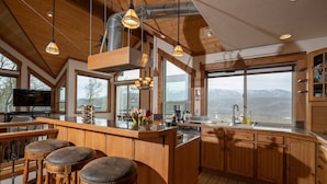 A spacious kitchen with wooden cabinets, a large island with stools, and a view of mountains through the windows. - Alpine Recharge (Alpine)