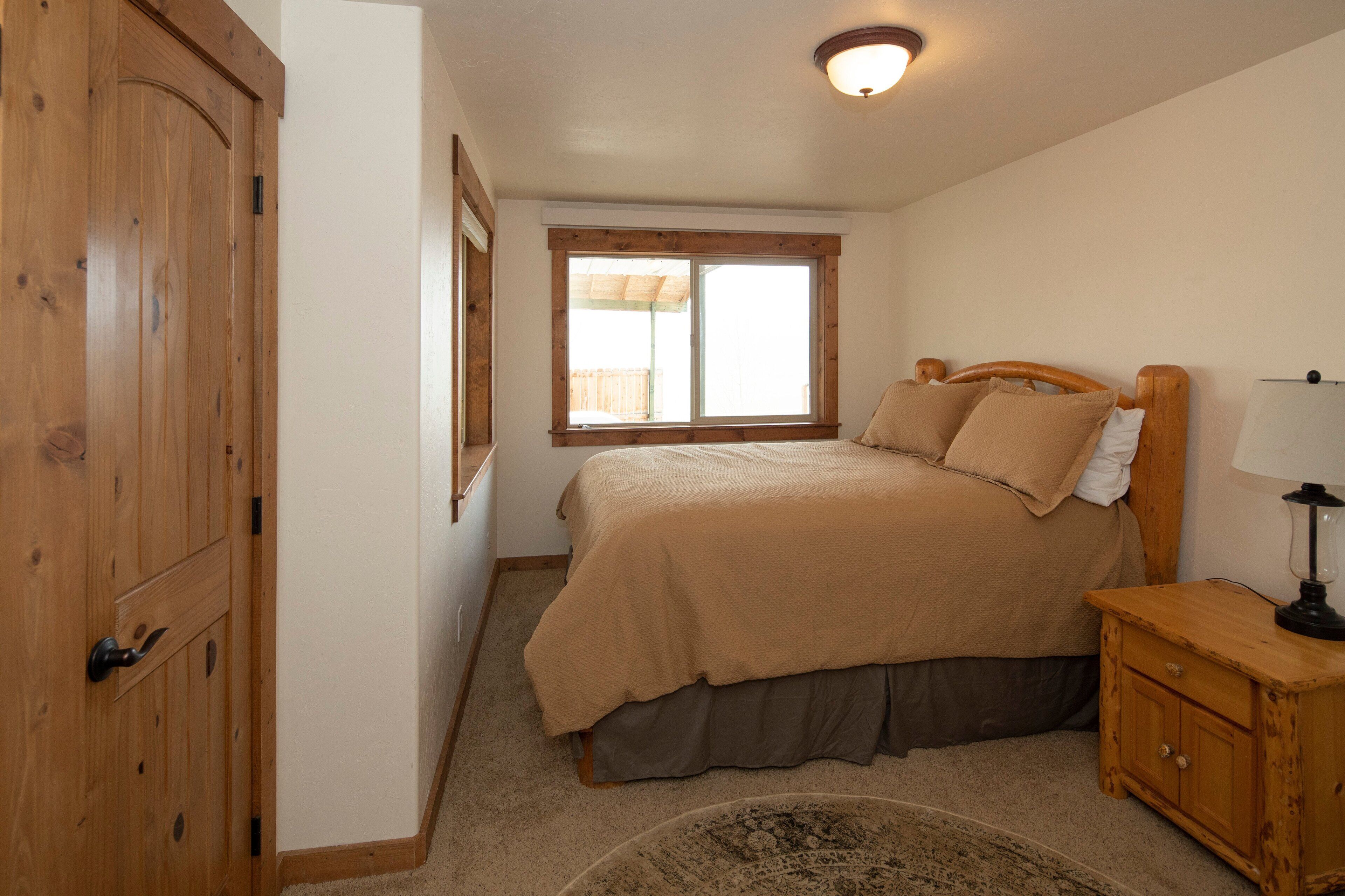 A bedroom with a wooden door, a bed with a beige bedspread, a nightstand with a lamp, and a window with a view of a fence.