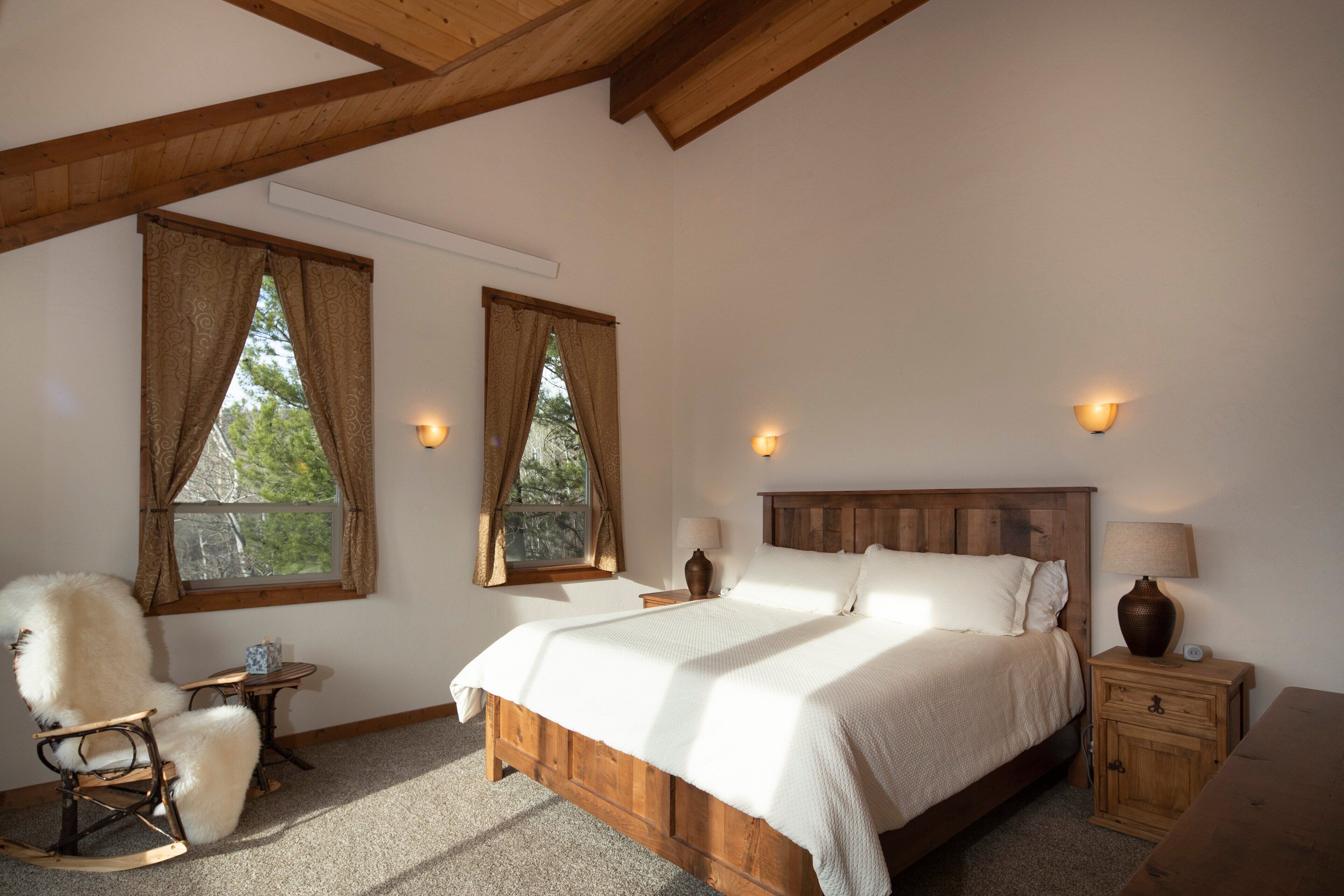 Une chambre à coucher avec un lit en bois, deux fenêtres avec des rideaux, un fauteuil à bascule, une table de chevet avec une lampe et une petite table.