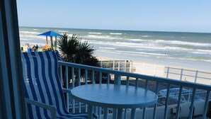 Een balkon met witte metalen hekwerk, een ronde tafel en gestreepte stoelen. In de verte de oceaan en een zandstrand.