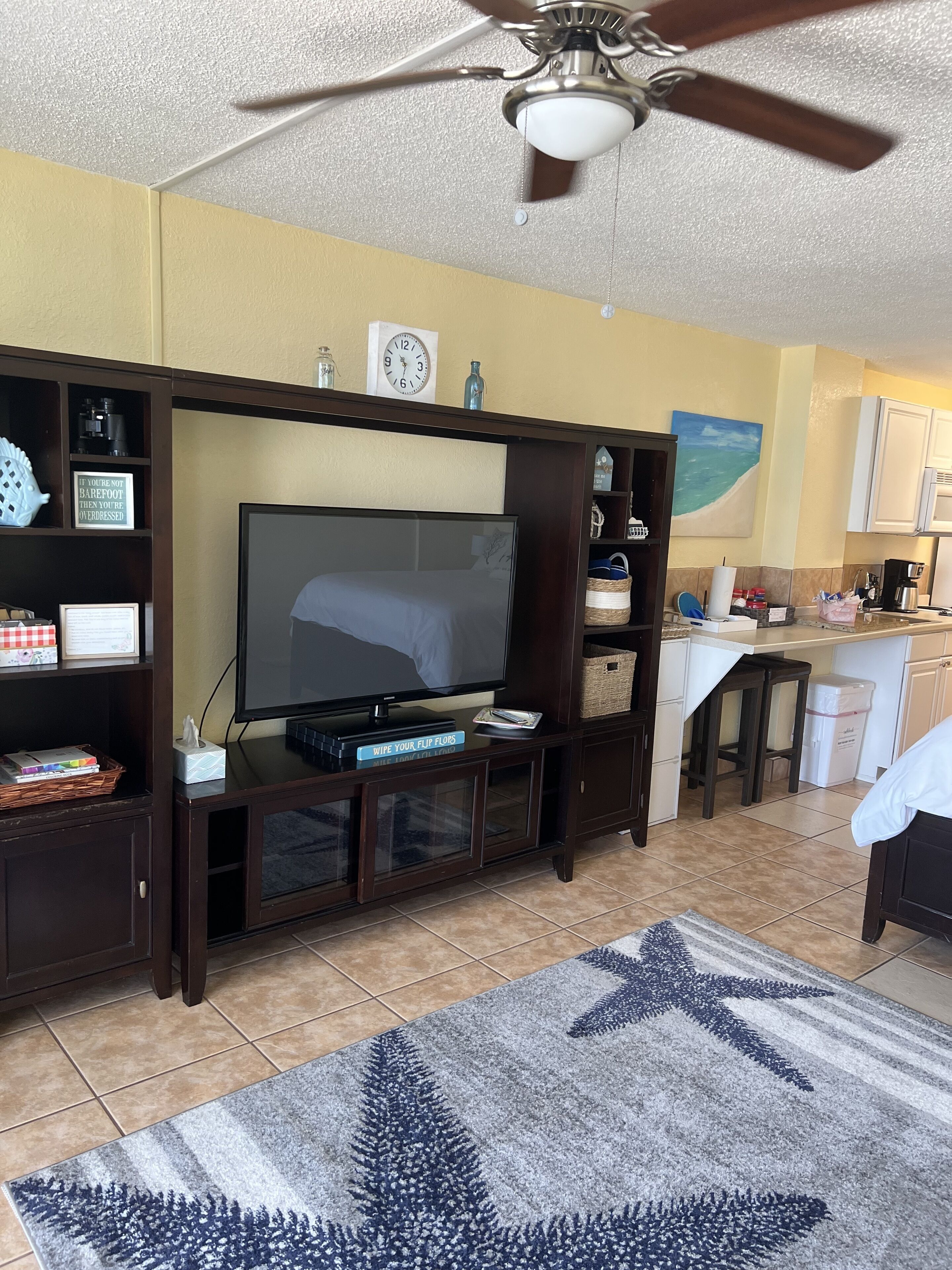 A living room with a TV, wooden entertainment center, kitchen area, and a ceiling fan.