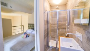A bedroom with a bed, a bedside table, and a nightstand. There is a shower area with a glass door and a white sink. - AF - Casa Rooms (Valletta)