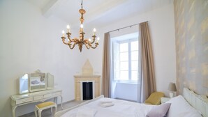 Un dormitorio con cama, tocador con espejo, candelabro, ventana con cortinas y mesita de noche con lámpara.