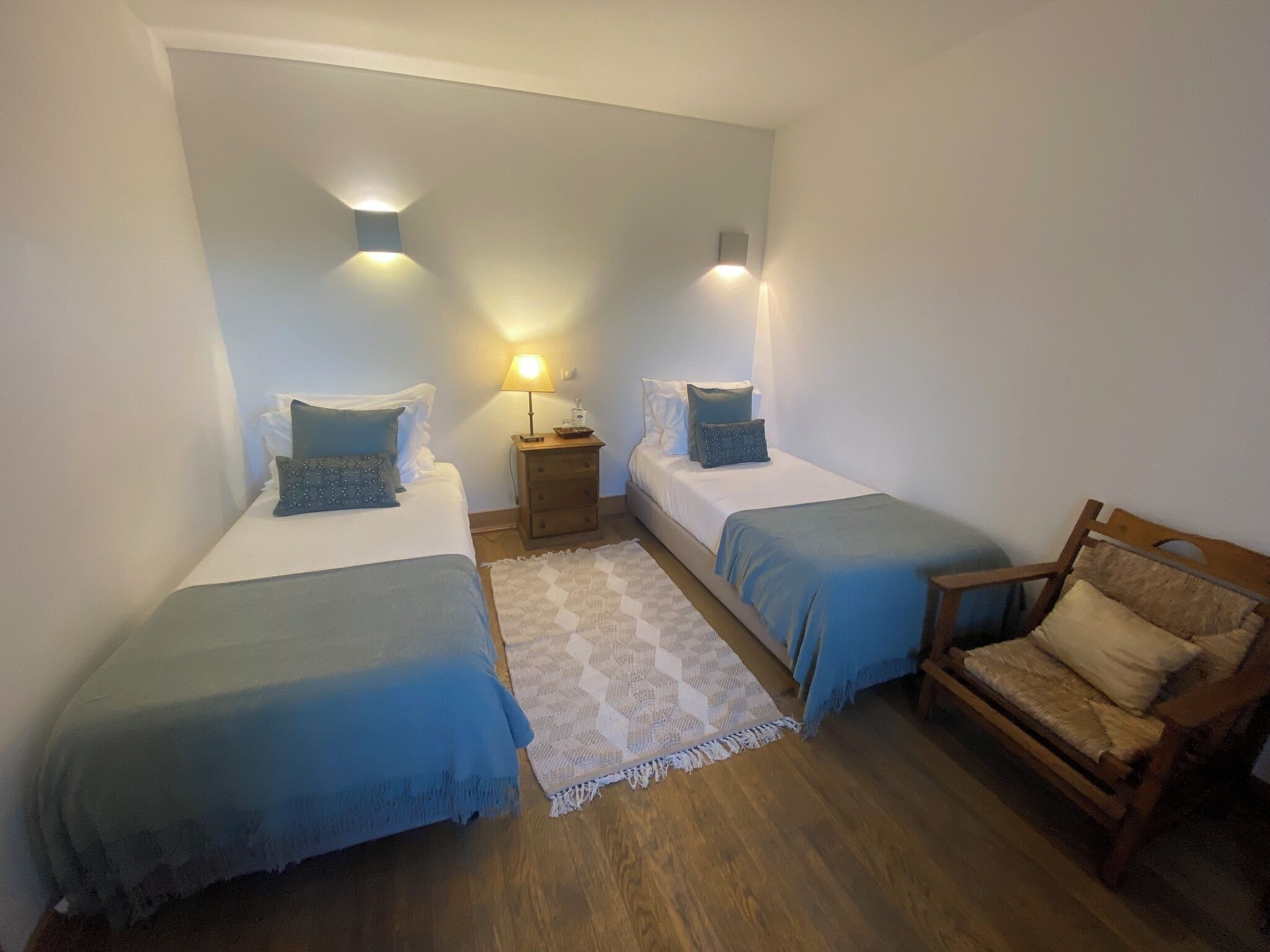 Een slaapkamer met twee eenpersoonsbedden, een houten stoel, een nachtkastje met een lamp en een kamerkleed met dessin.
