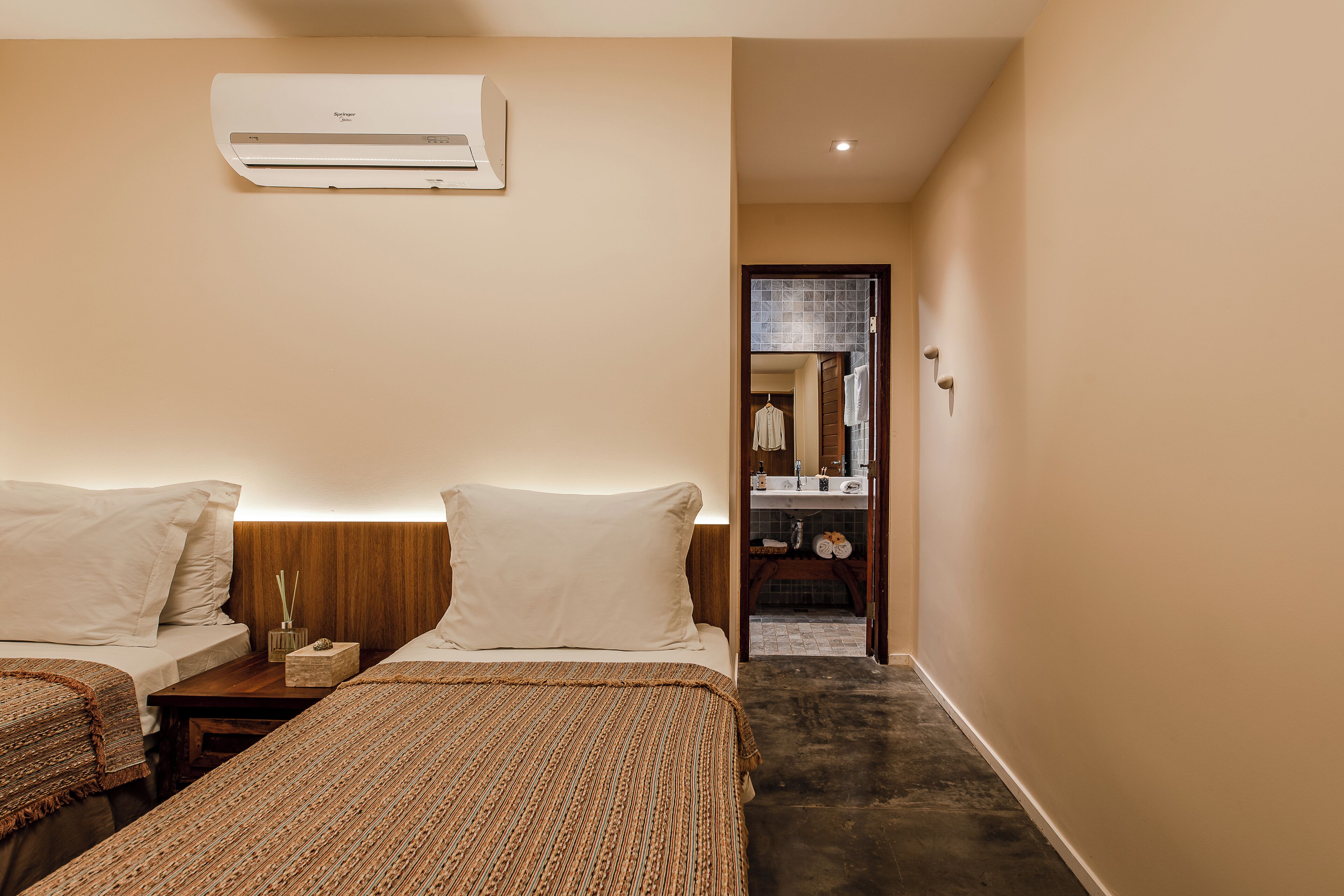 A hotel room with two beds, a wooden nightstand, a wall-mounted air conditioner, and a bathroom visible through an open door.
