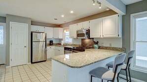 A modern kitchen with white cabinets, a granite countertop, stainless steel appliances, and a large island with bar stools. - Ocean Retreat with Private Beach Access (Kure Beach)