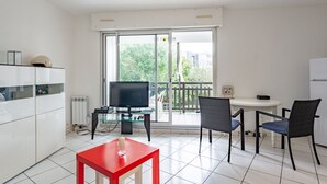 A modern kitchen with a TV, dining table, and chairs, a red table in the foreground, and a sliding glass door to a balcony.