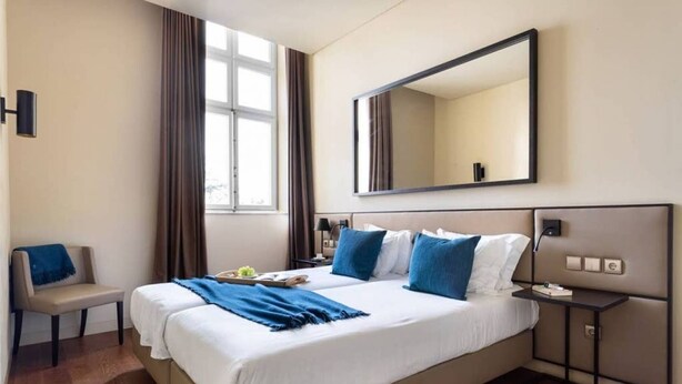 A hotel room with a large bed, a bedside table, a chair, a window with curtains, and a wall-mounted mirror. - JOIVY Urban 2BR Apt w/ Park view, in Cais do Sodré (Lisboa)