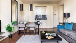 A modern living room with a sofa, coffee table, TV, and a dining area with chairs and a kitchen in the background. - JOIVY Urban 2BR Apt w/ Park view, in Cais do Sodré (Lisboa)