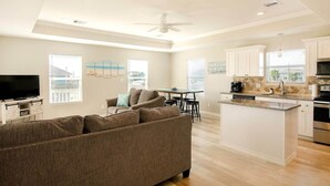 Ein modernes Wohnzimmer mit einer großen Couch, einem Essbereich mit Tisch und Stühlen, einer Küche mit weißen Schränken und einer Kochinsel sowie einem Deckenventilator.