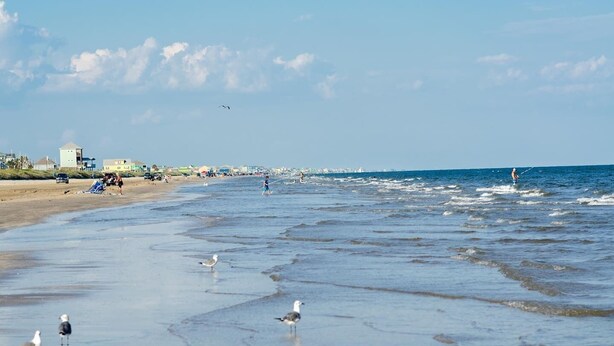 Ein Strand mit Wellen, Menschen und Gebäuden im Hintergrund.