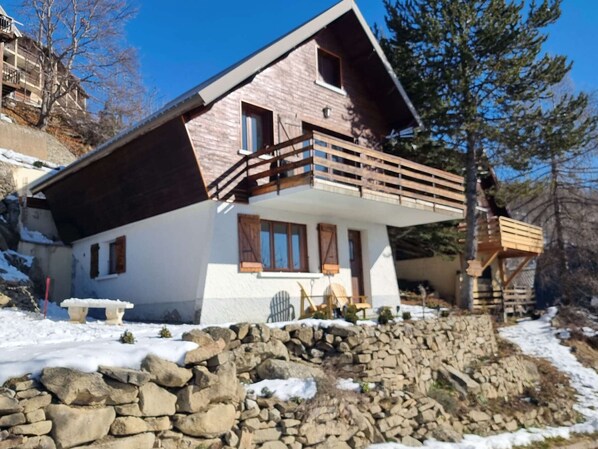 A two-story house with a stone foundation, wooden balcony, and a stone wall in front.