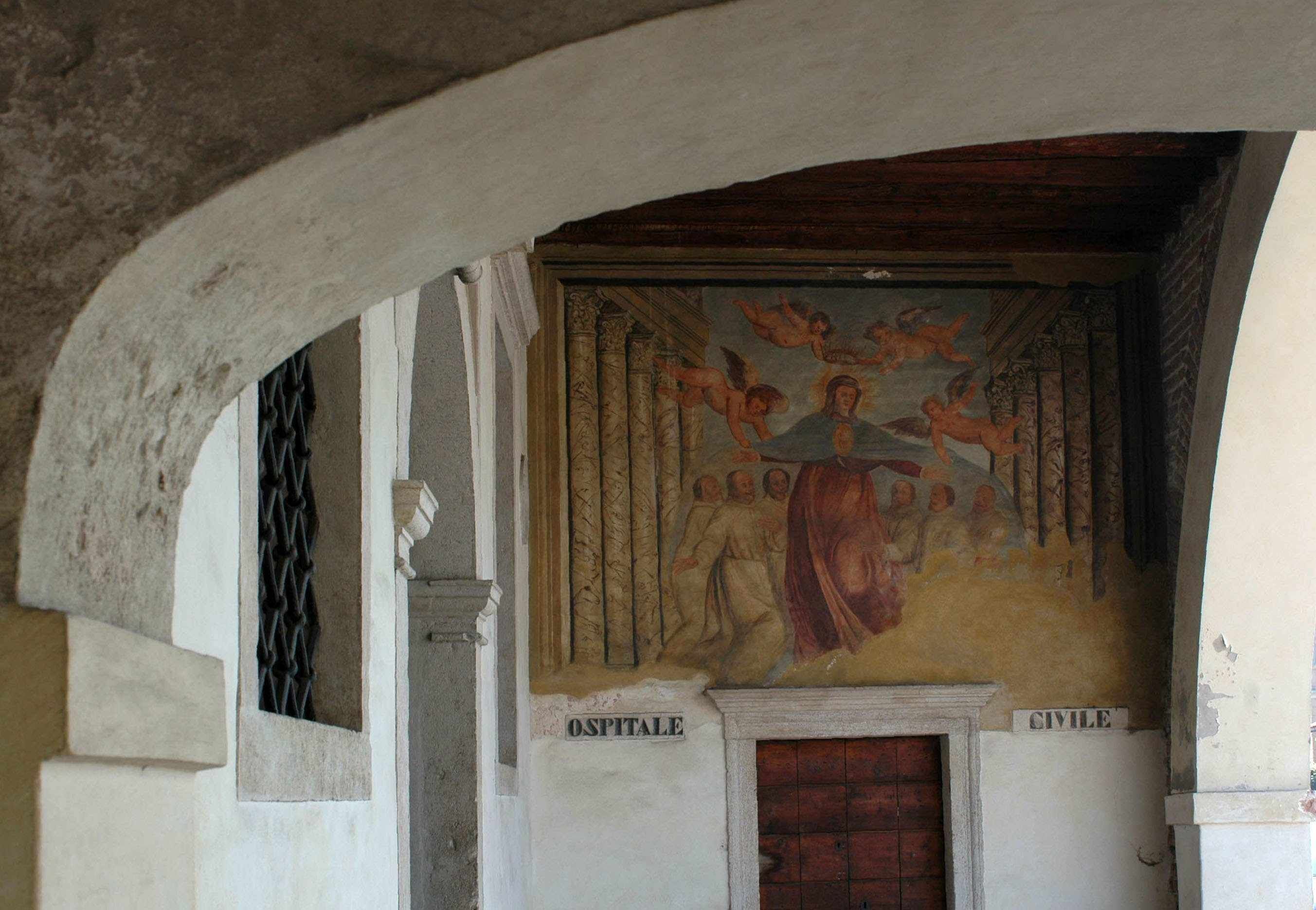 A historical building interior with a fresco mural, a wooden door, and two columns labeled "OSPITALE" and "CIVILE".