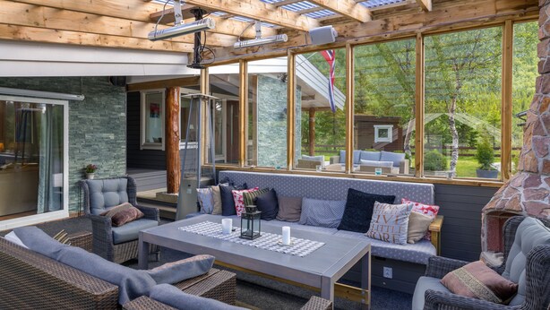 An outdoor living area with wicker furniture, a table with candles, and large windows offering a view of the surrounding greenery. - VESTERALEN LODGE - TOP QUALITY IN THE VESTERALEN and LOFOTEN (Sortland)