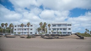 Ein Hotel direkt am Strand mit mehreren Etagen, Palmen und einem sandigen Strand im Vordergrund.