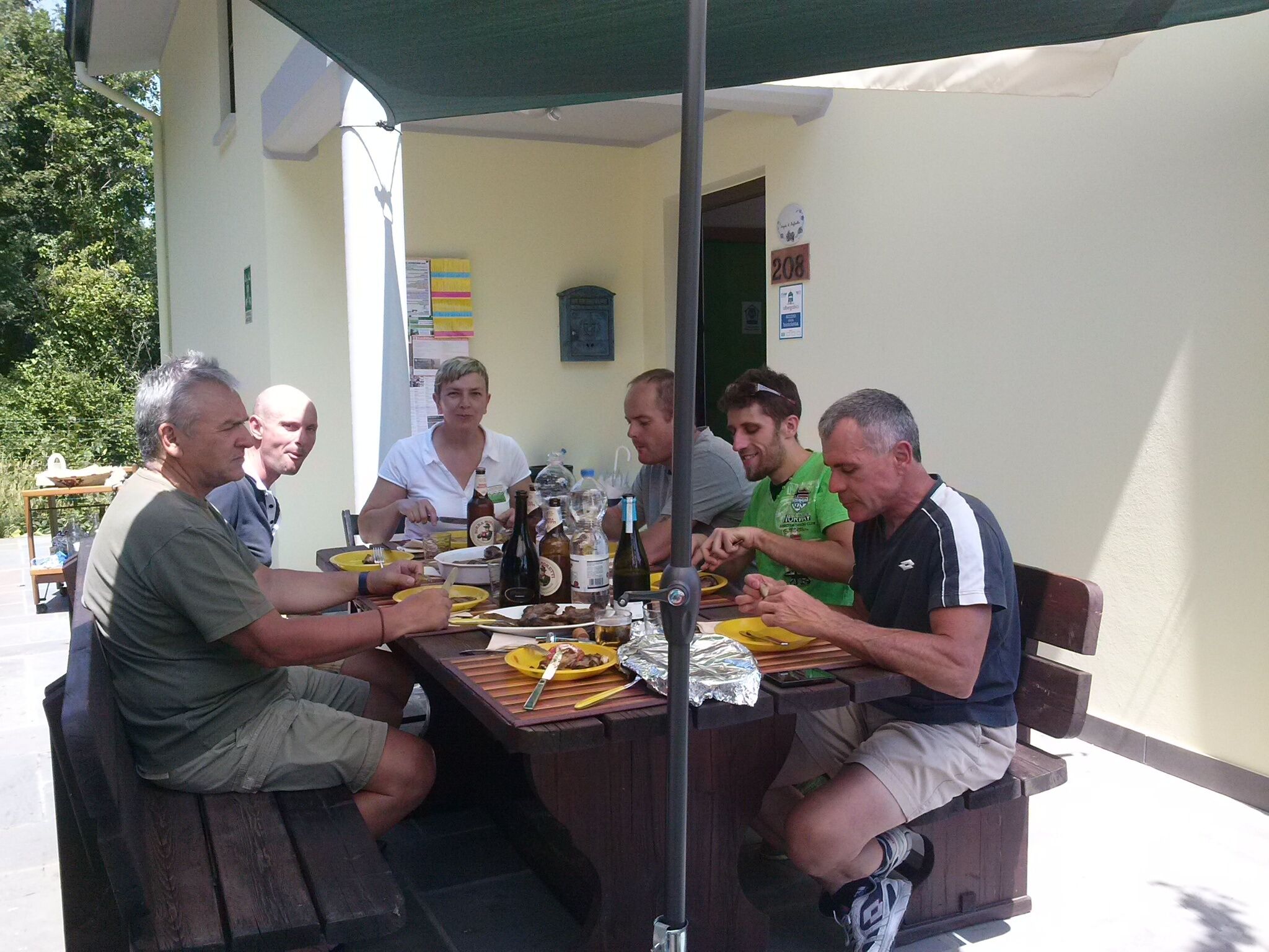 A group of people dining at an outdoor table with a green umbrella, surrounded by a white building with a door marked '208'.