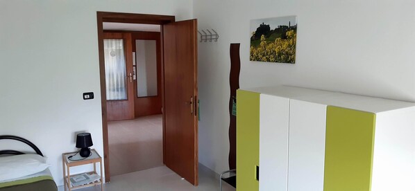 Una camera d'albergo con un letto, un tavolino con una lampada, un armadio e una porta che dà su un'altra stanza.