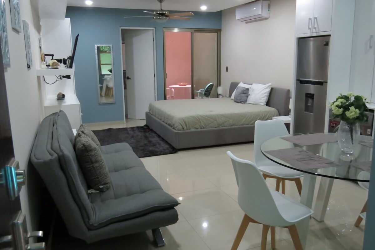 Una habitación de hotel compacta con cama, sofá, mesa de comedor con sillas, ventilador de techo y aire acondicionado.