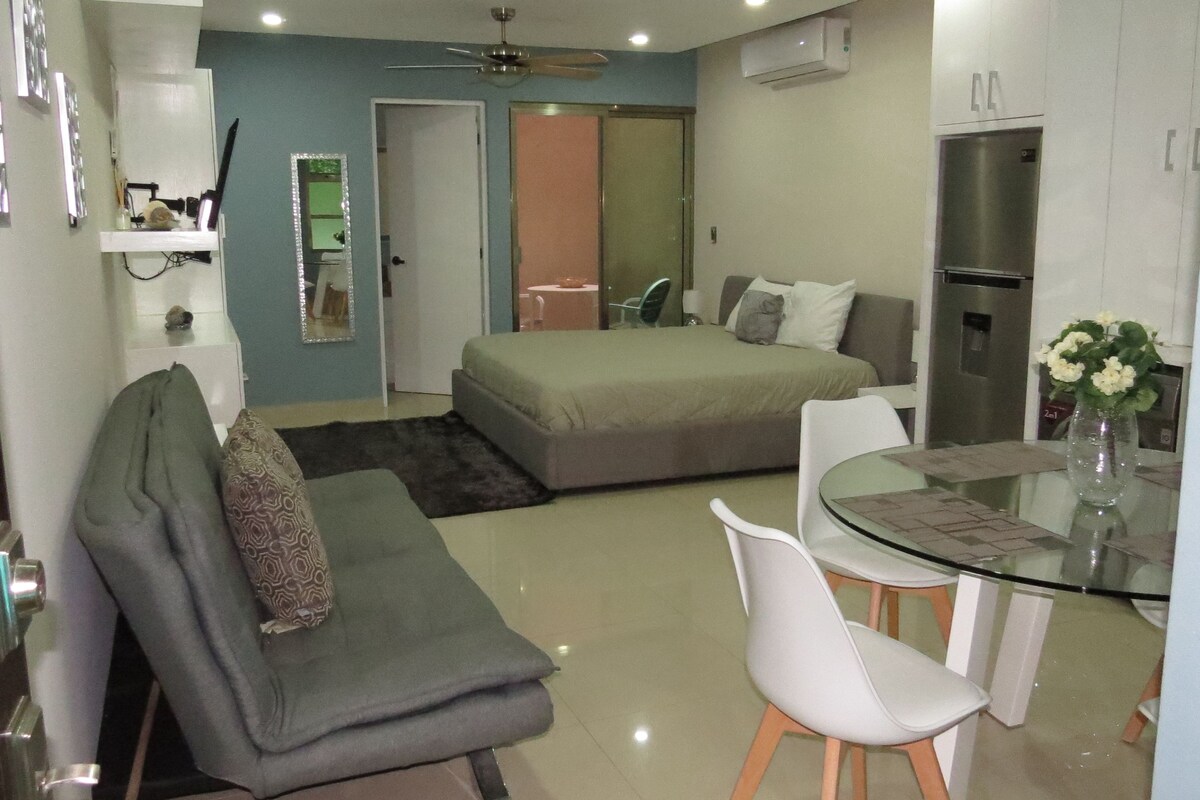 A compact living space with a bed, a sofa, a dining table with chairs, a ceiling fan, and a refrigerator.