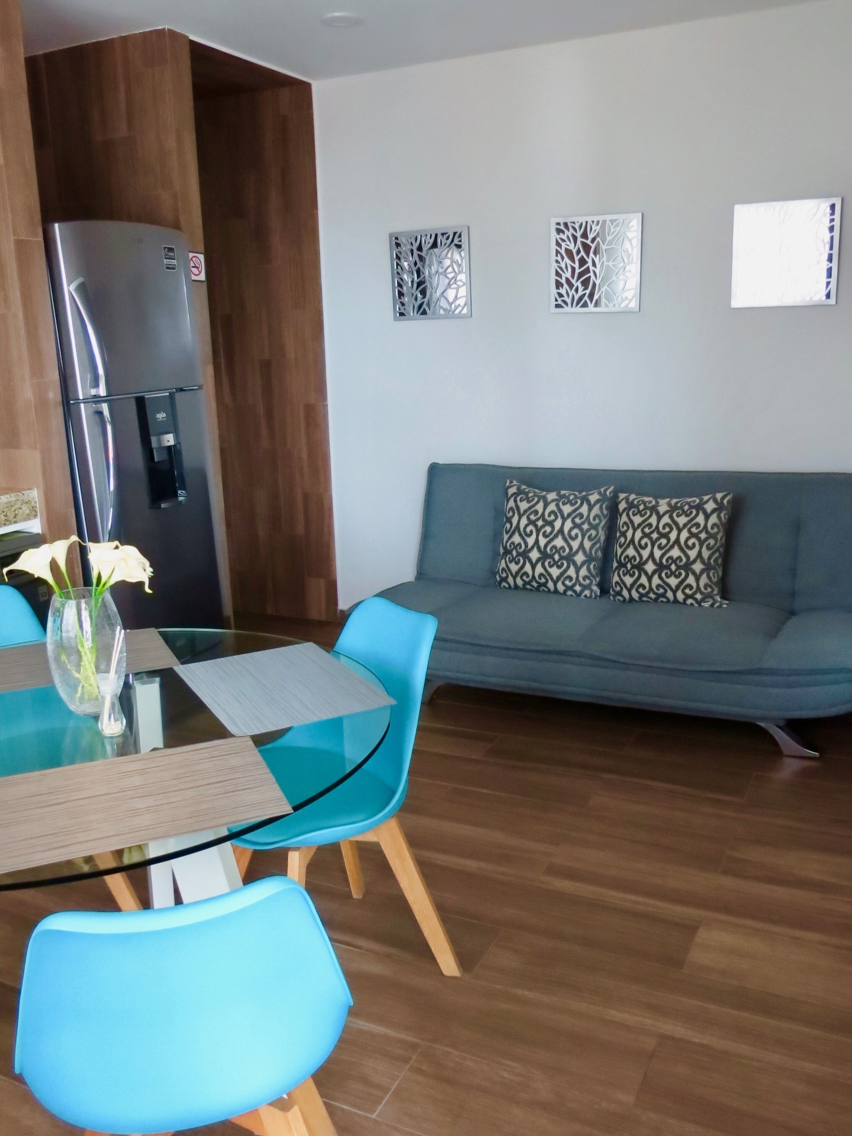 A modern living room with a glass-top dining table, blue chairs, a grey sofa with patterned cushions, a built-in wooden cabinet, and a stainless steel refrigerator.