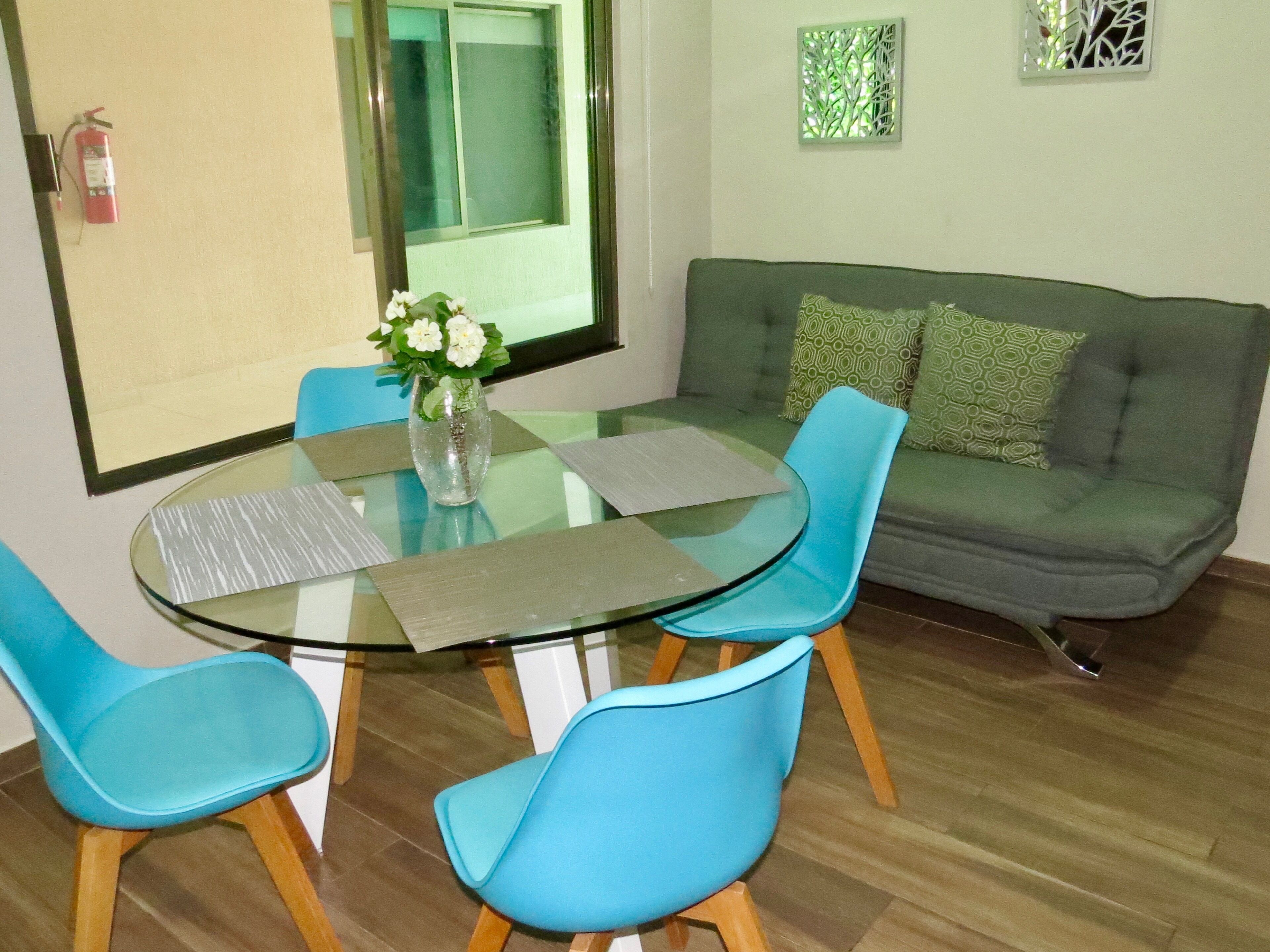 A dining area with a glass table, blue chairs, a vase with flowers, a green sofa with patterned cushions, and a fire extinguisher on the wall.